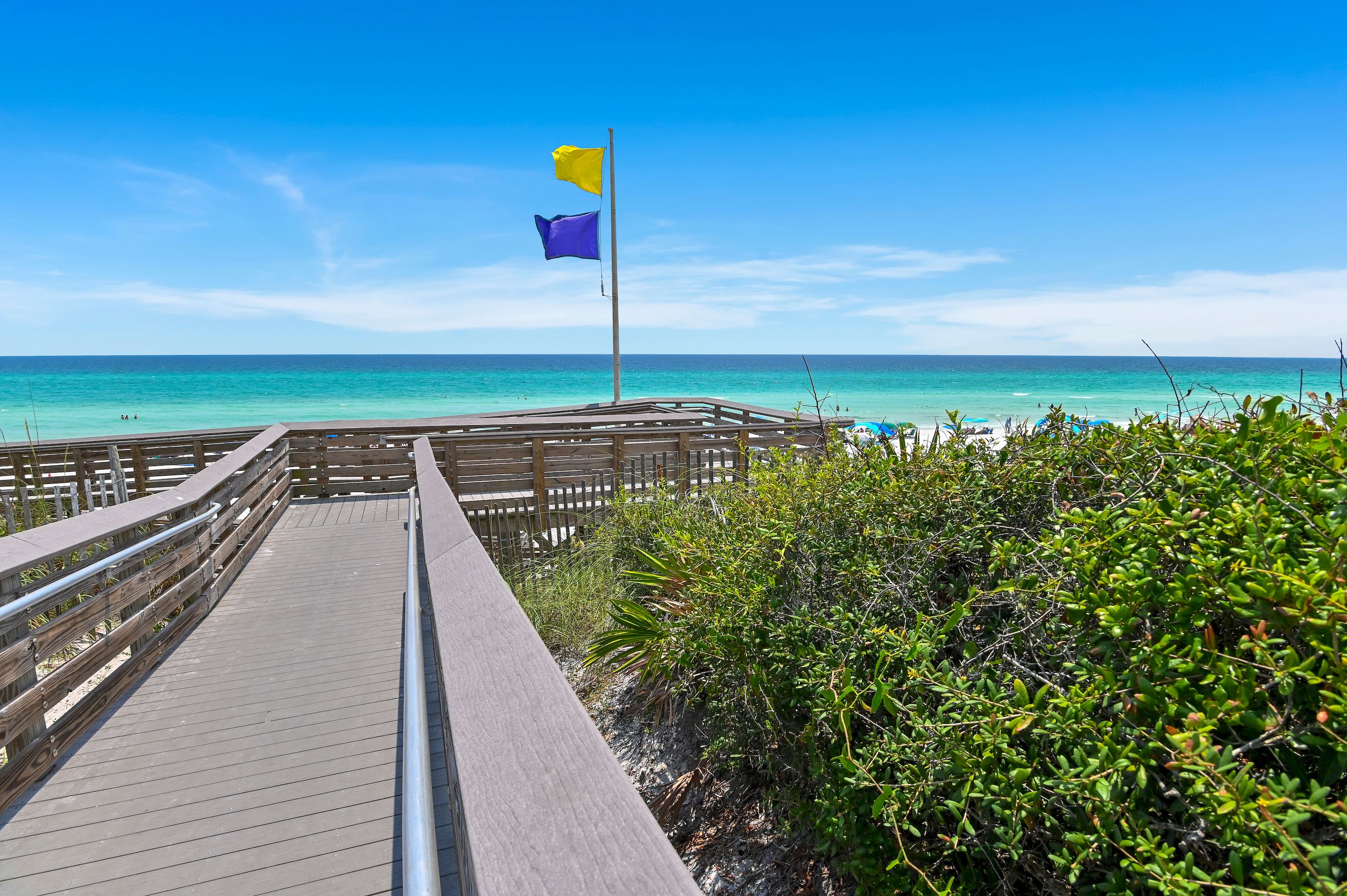 A wooden boardwalk leading to a beach with clear turquoise water, a flagpole with a yellow and purple flag, and lush greenery.