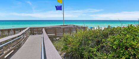 A wooden boardwalk leading to a beach with clear turquoise water, a flagpole with a yellow and purple flag, and lush greenery.