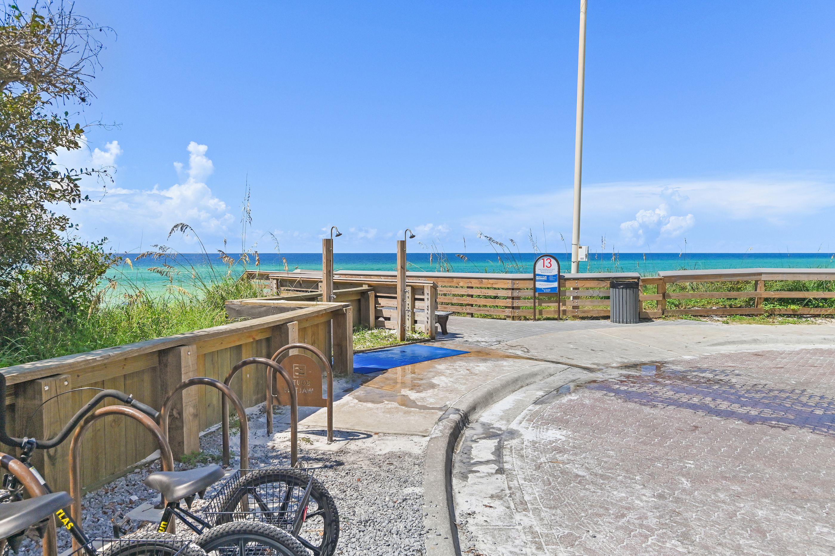 A wooden pathway leading to a beach with a clear view of the ocean, a signpost, and a bicycle parked on the side.