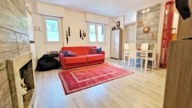 A living room with a red sofa, a fireplace, a dining area with a table and chairs, and a kitchen area with cabinets. - Elegant Apartment in the center of Aprica, very close to the ski slopes (Aprica)