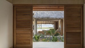 A bedroom with a view of a beach, featuring wooden shutters, a thatched roof, and a bed.