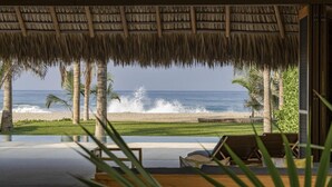 A beach view from a room with a thatched roof, palm trees, and a pool.