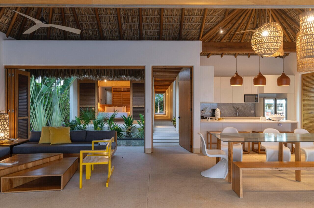 A modern interior with a thatched ceiling, a dining area with white chairs, a kitchen with stainless steel appliances, and a living area with a sofa and yellow chairs.