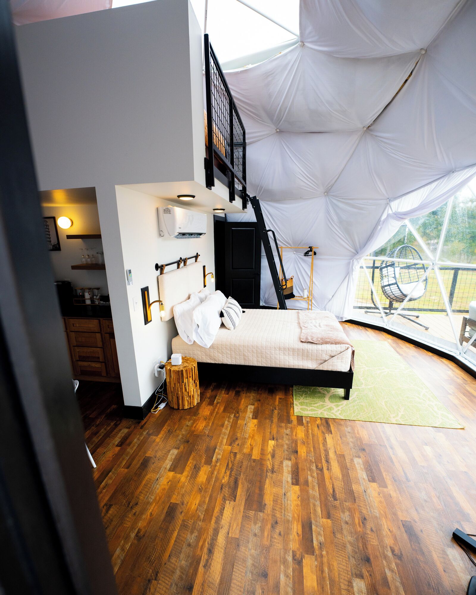 A bedroom with a bed, wooden flooring, a hanging chair, and a view of the outdoors.