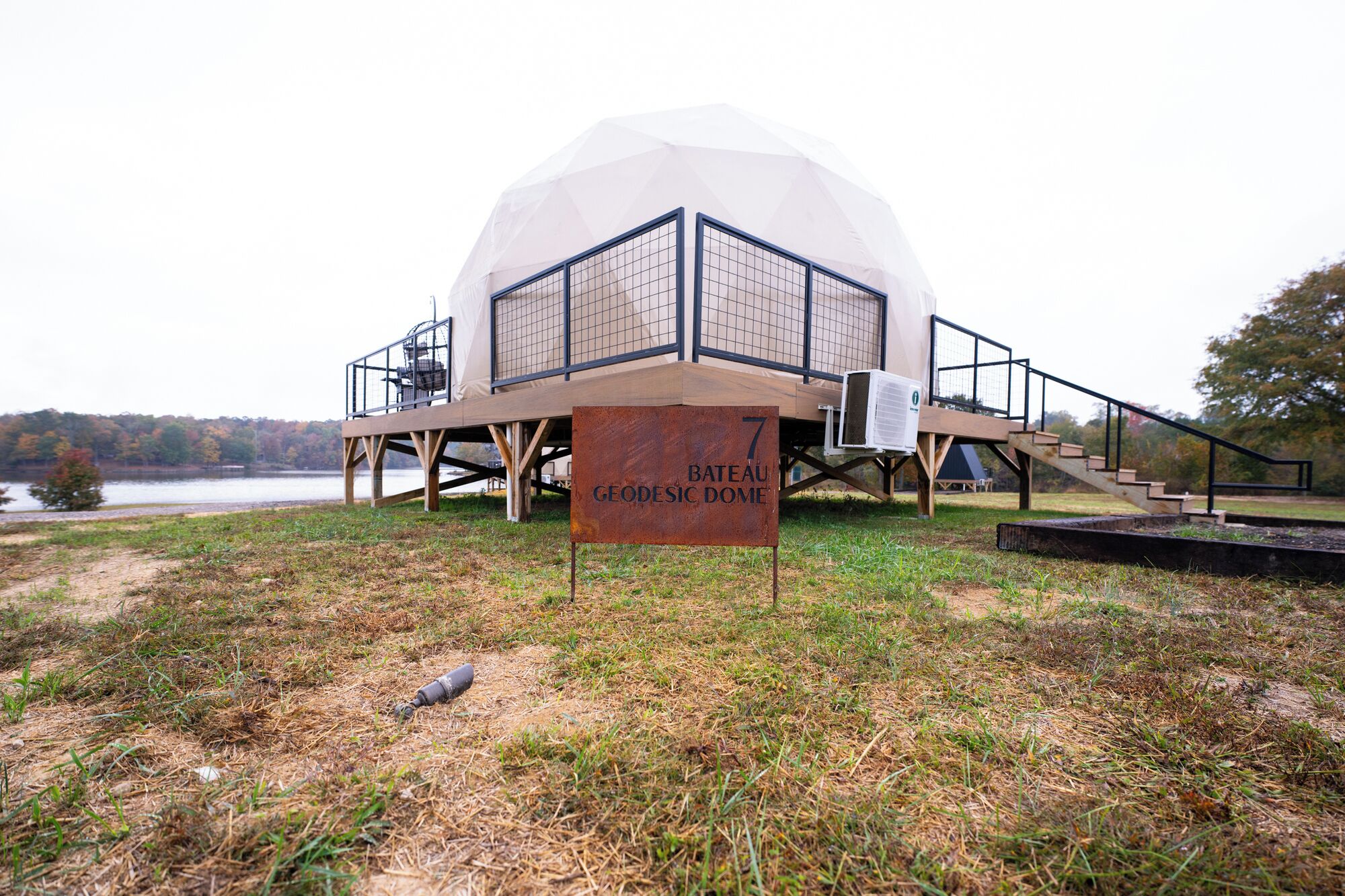 Uma estrutura em forma de cúpula geodésica com uma placa indicando ser o 7º edifício, chamado Cúpula Geodésica Bateau, situado perto de um corpo d'água com um gramado à frente.