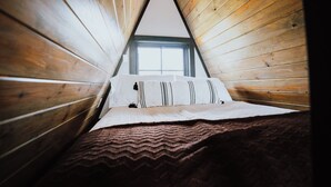 A triangular attic bedroom with wooden walls, a window, and a bed with patterned bedding. - Modern A-Frame Cabin: Hot Tub, TV, & Lake Views (Cedar Bluff)
