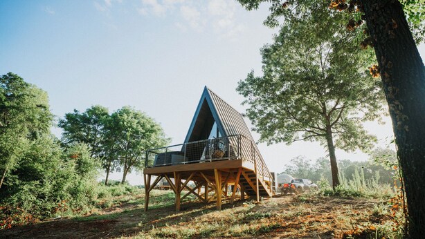 A modern, triangular wooden structure with a glass balcony, surrounded by trees and natural landscape. - Modern A-Frame Cabin: Hot Tub, TV, & Lake Views (Cedar Bluff)
