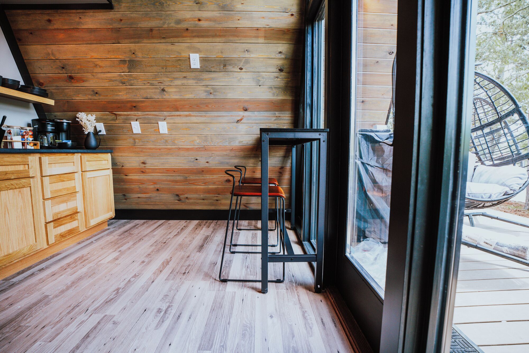 A kitchen with wooden walls, a black metal stool, and a window overlooking a deck.