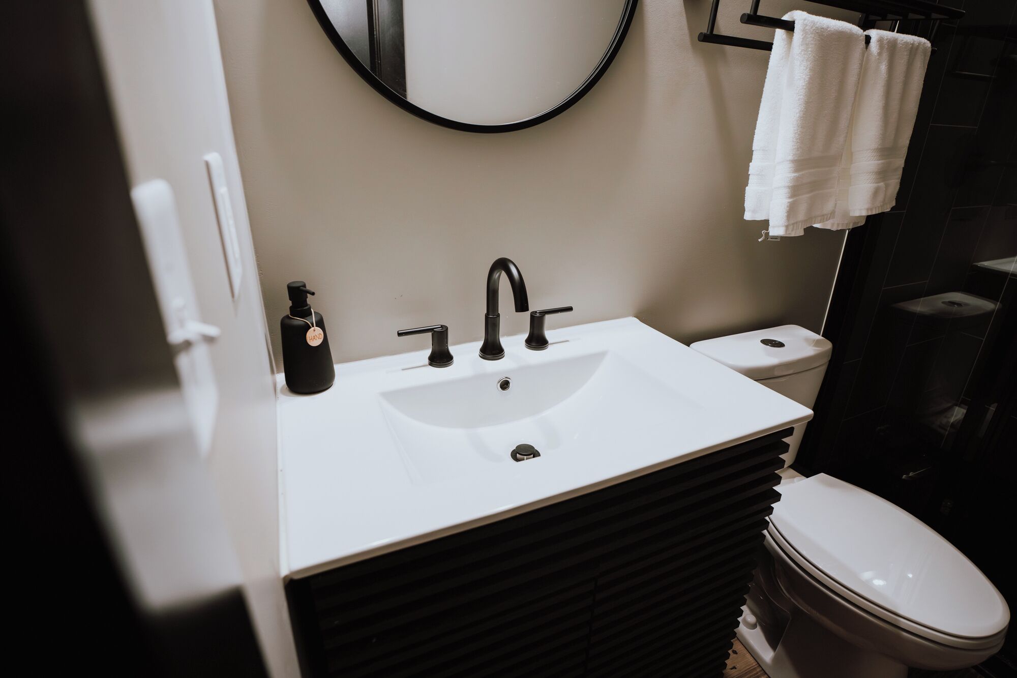 Un baño moderno con lavamanos blanco, grifo negro y un inodoro. Encima del lavamanos hay un espejo redondo y un perchero con toallas.