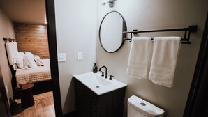 A compact bathroom with a white sink, black faucet, and a toilet. There is a wooden bench with a white towel and a pillow, and a round mirror on the wall. - Lake Front Modern Cabin: Hot Tub, TV, & Lake Views (Cedar Bluff)