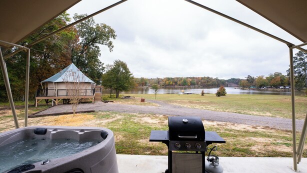 A covered outdoor area with a hot tub, grill, and a view of a lake and trees. - Lake Front Luxury Glamping Tent Suite: Hot Tub & Lake Views (Cedar Bluff)