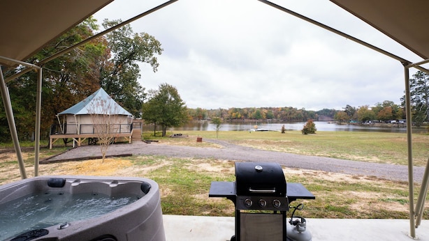 Een overdekt buitengebied met een jacuzzi, barbecue en uitzicht op een meer en bomen.