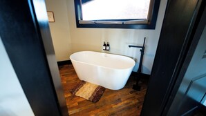 A modern bathroom with a freestanding bathtub, wooden flooring, and a window above the sink.