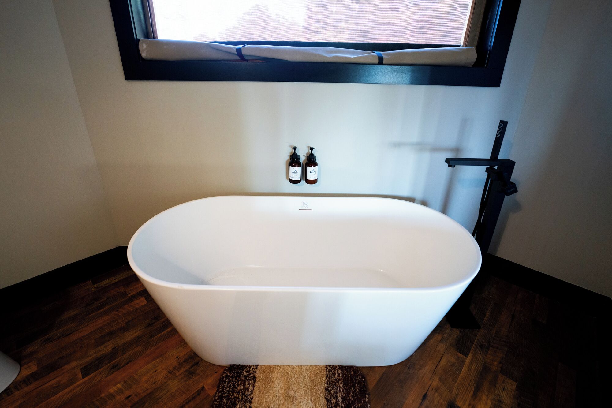 A modern bathroom with a freestanding bathtub, a window above the sink, and two bottles of hand soap on the wall.