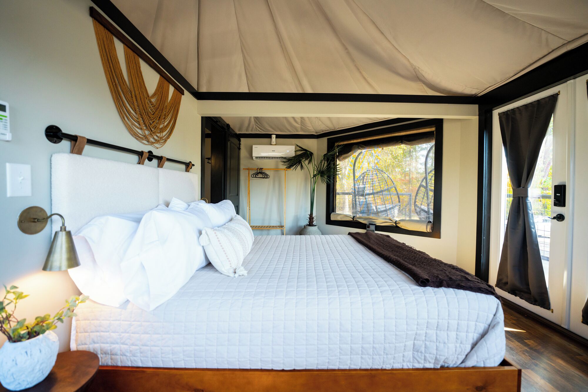 Un'ampia camera da letto con un letto grande, un comodino con una lampada, un decoro in macramè e vista sull'esterno da una grande finestra.