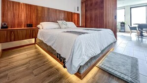 Ein modernes Schlafzimmer mit einem Holz-Kopfteil, einem großen Bett mit weißer Bettwäsche, einem Deckenventilator und Blick auf den Wohnbereich durch eine Glastür.