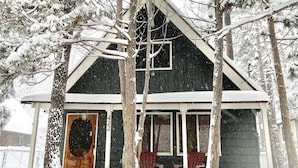 Une cabane en bois recouverte de neige, avec un porche à l’avant, une grande fenêtre et une porte.