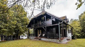 Une maison en bois à deux étages, dotée d’un balcon, entourée d’arbres et bordée d’une pelouse.