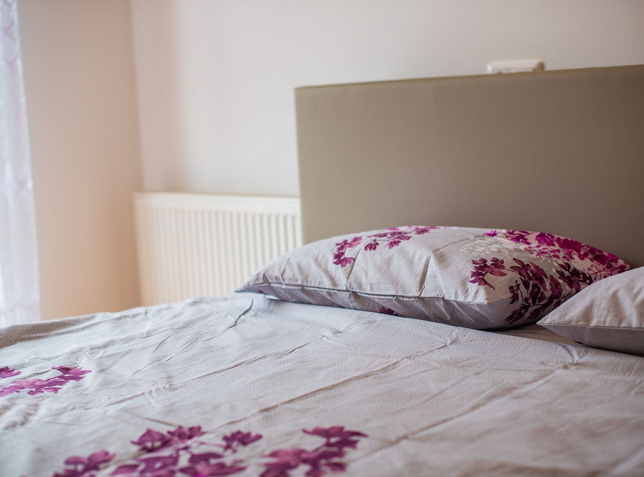 Una cama con cubrecama y almohadas de estampado floral, un cabecero sencillo y un radiador blanco al fondo.