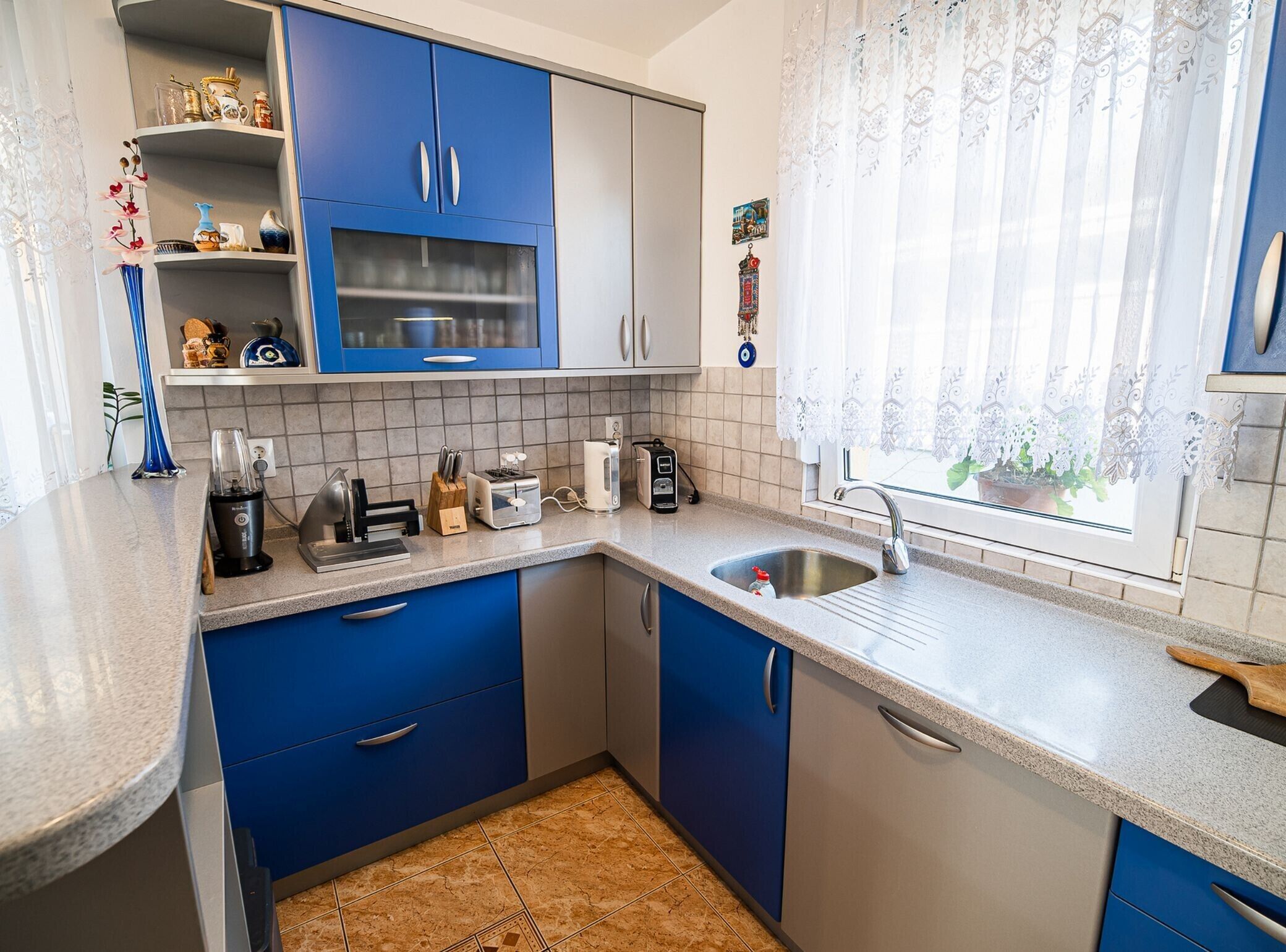 A compact kitchen with blue and white cabinets, a granite countertop, and a window with lace curtains.