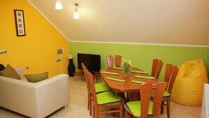 Uma área de refeições com uma mesa e cadeiras de madeira, um sofá, uma televisão e um puff amarelo.