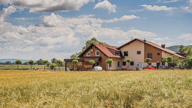 Uma casa de dois andares com telhado vermelho, rodeada por um campo de trigo, e um céu claro com nuvens.