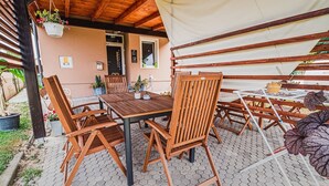 Uma área de pátio externa com mesa e cadeiras de madeira, plantas em vasos e um espaço de estar coberto.