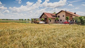 A two-story building with a red-tiled roof, surrounded by a field of crops, and parked cars in the driveway.