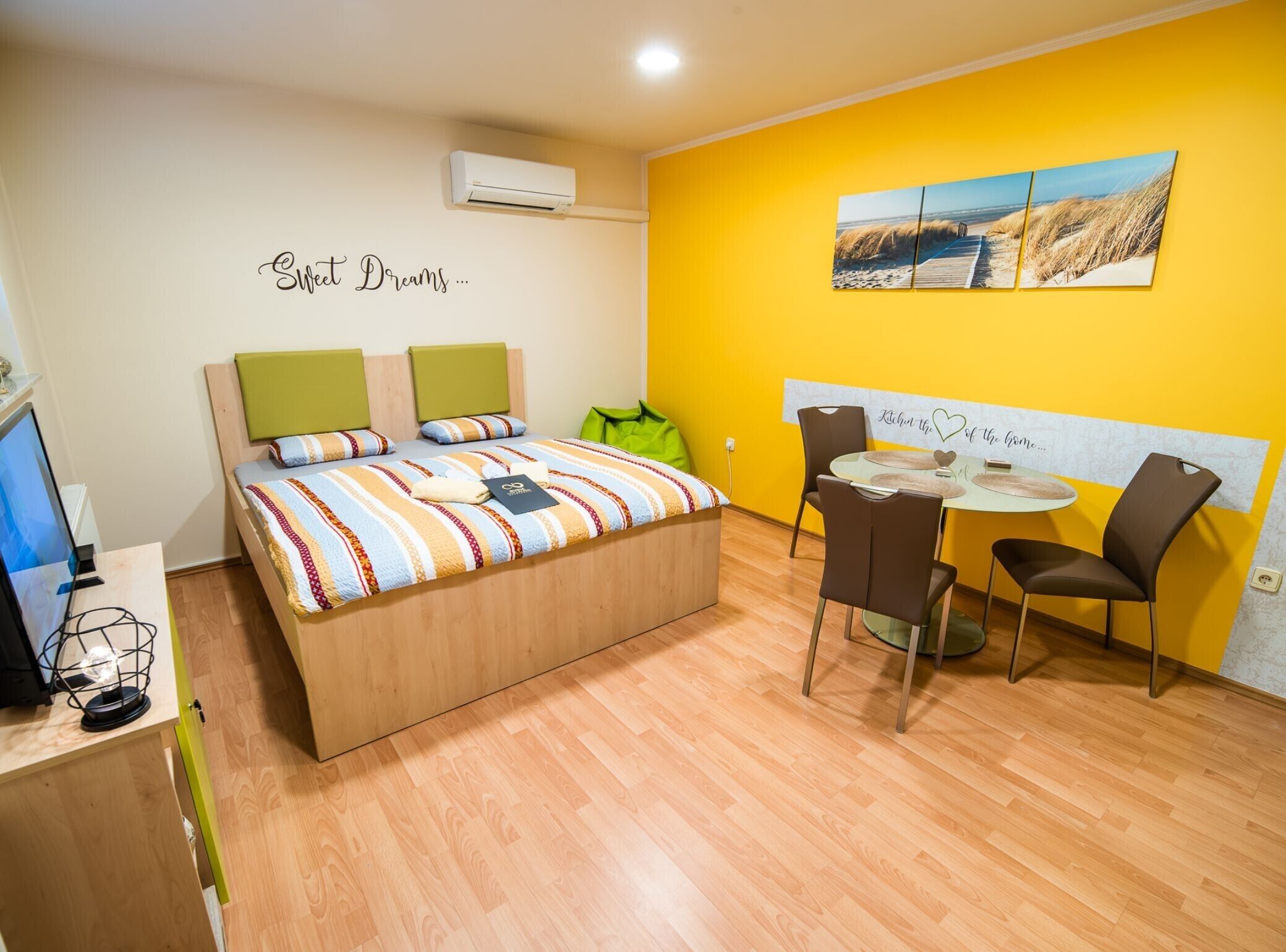 A bedroom with a bed, bedside table, and a desk with a computer. There is a window, a chair, and a table with a painting on the wall.