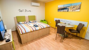A bedroom with a bed, bedside table, and a desk with a computer. There is a window, a chair, and a table with a painting on the wall.
