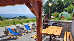 Een terras buiten met houten meubels, waaronder een tafel en stoelen, en een barbecue.