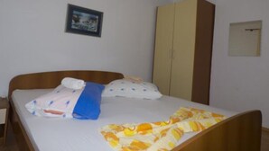 Een enkel bed met een deken met witte en gele strepen, een blauw kussen en een wit kussen. Rechts staat een houten garderobe en erboven, aan de muur, hangt een lijst met een foto.