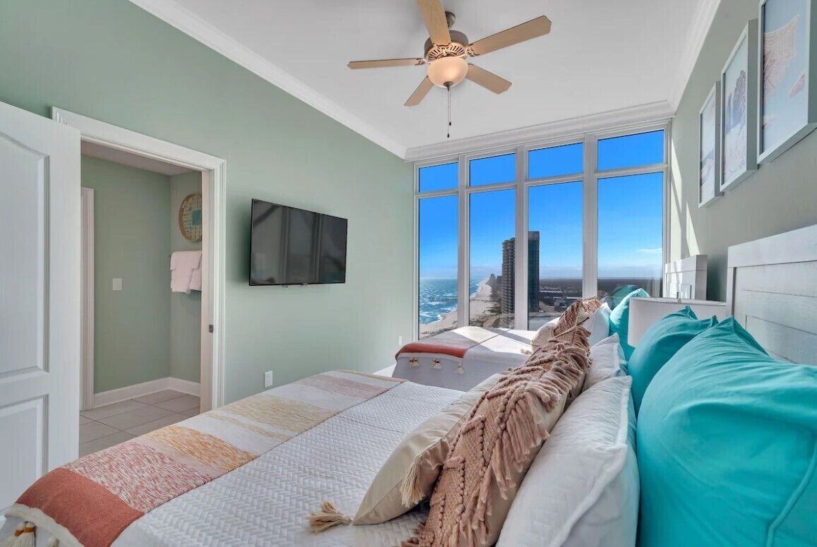 A bedroom with a large bed, a ceiling fan, a TV mounted on the wall, and a view of the ocean through large windows.