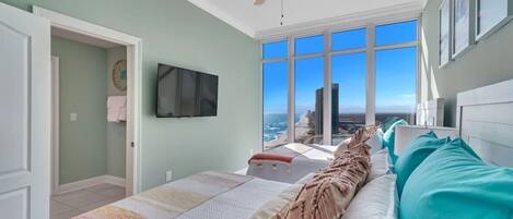 A bedroom with a large bed, a ceiling fan, a TV mounted on the wall, and a view of the ocean through large windows.
