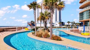 A pool area with palm trees, lounge chairs, and a building with a modern design. - Luxury 4BR Beachfront Corner Unit, Panoramic Gulf Views, Lazy River, Indoor Pool (Orange Beach)