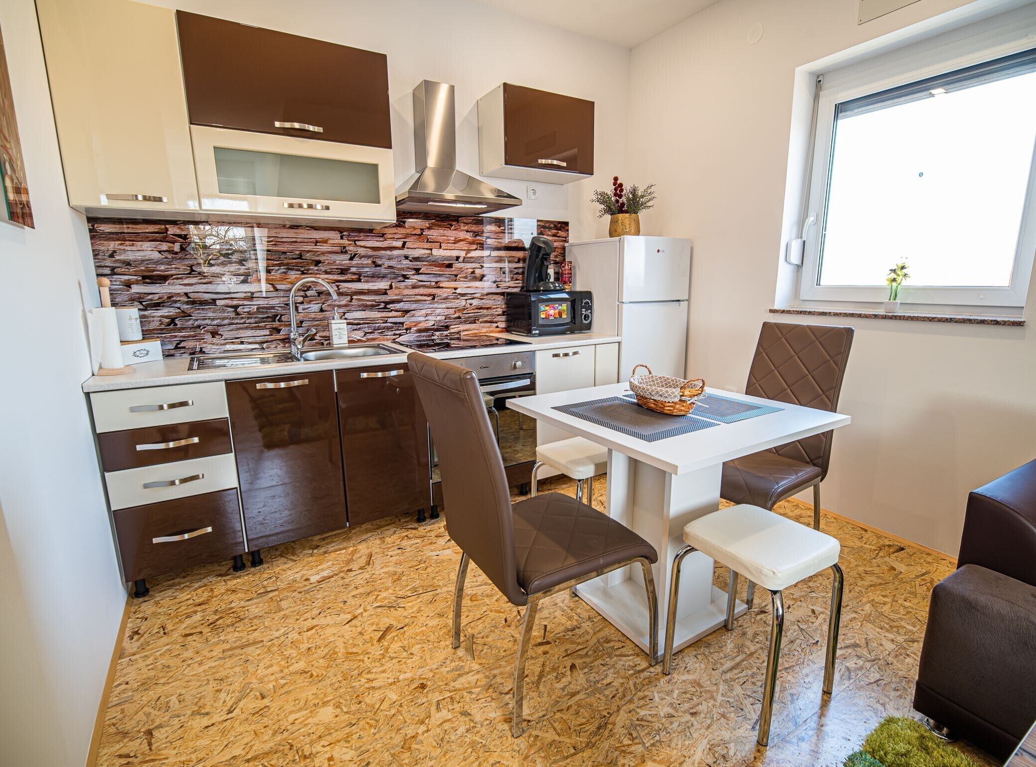A compact kitchen with a stone backsplash, a microwave, a stove, and a refrigerator. There is a dining table with two chairs and a small window.