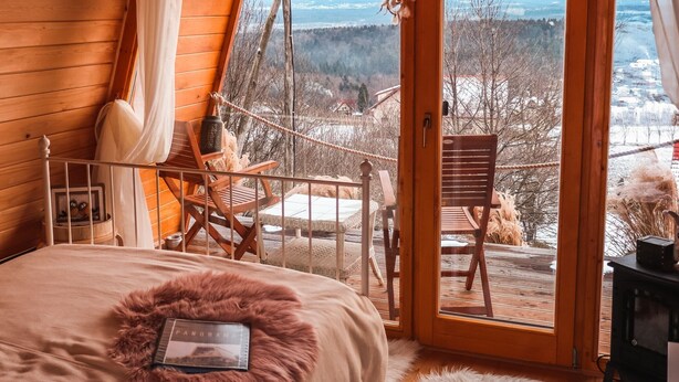 A cozy wooden cabin room with a bed, a small table, and a chair. There is a balcony with a view of a snowy landscape and mountains.