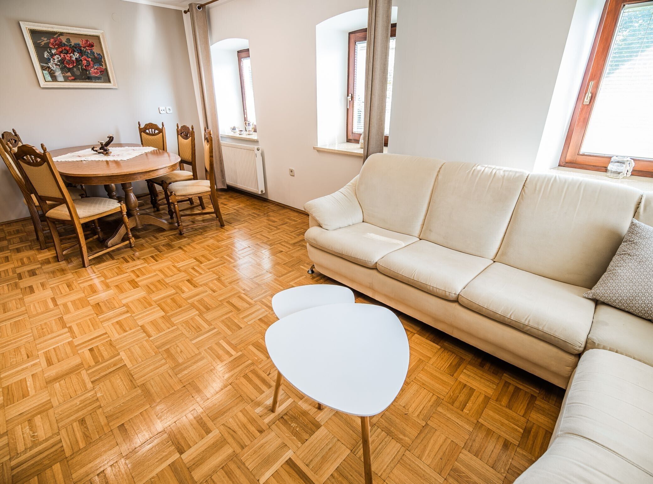 Ein Wohnzimmer mit Holzboden, einem beigefarbenen Sofa, einem kleinen weißen Tisch und einem Esstisch mit Stühlen.