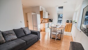 A modern living room with a grey sofa, wooden flooring, a dining area with a wooden table and chairs, a kitchen with white cabinets, and a large window with a view of a snowy landscape.