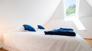 Camera da letto con un letto, biancheria bianca, cuscini blu e una coperta blu, con una finestra che si affaccia su una montagna.