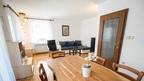 Zona pranzo con tavolo e sedie in legno, un divano, un termosifone e una televisione sullo sfondo.
