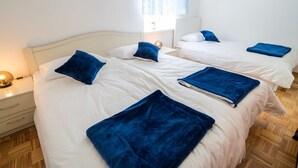 Camera d'albergo con due letti, ciascuno con biancheria da letto bianca e cuscini blu. C'è un comodino con una lampada e un cassetto.