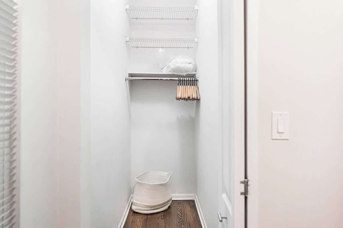 A narrow hallway with white walls, a wooden floor, and a white door. There are wire shelves with a white bag and a basket on the top shelf, and a wooden rack with handles on the middle shelf.