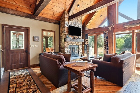 A rustic interior with a stone fireplace, wooden beams, and a large window overlooking greenery. - Big Woods and Yellow River (Necedah)