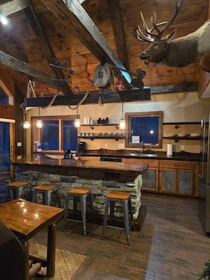 A rustic kitchen with a stone island, wooden bar stools, and a mounted elk head.