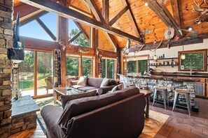 A rustic interior with a stone fireplace, wooden beams, a stone bar, and a large window overlooking a natural landscape.