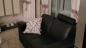 Ein modernes Wohnzimmer mit einem schwarzen Ledersofa, einem gemusterten Zierkissen und einem Fenster mit Vorhängen.
