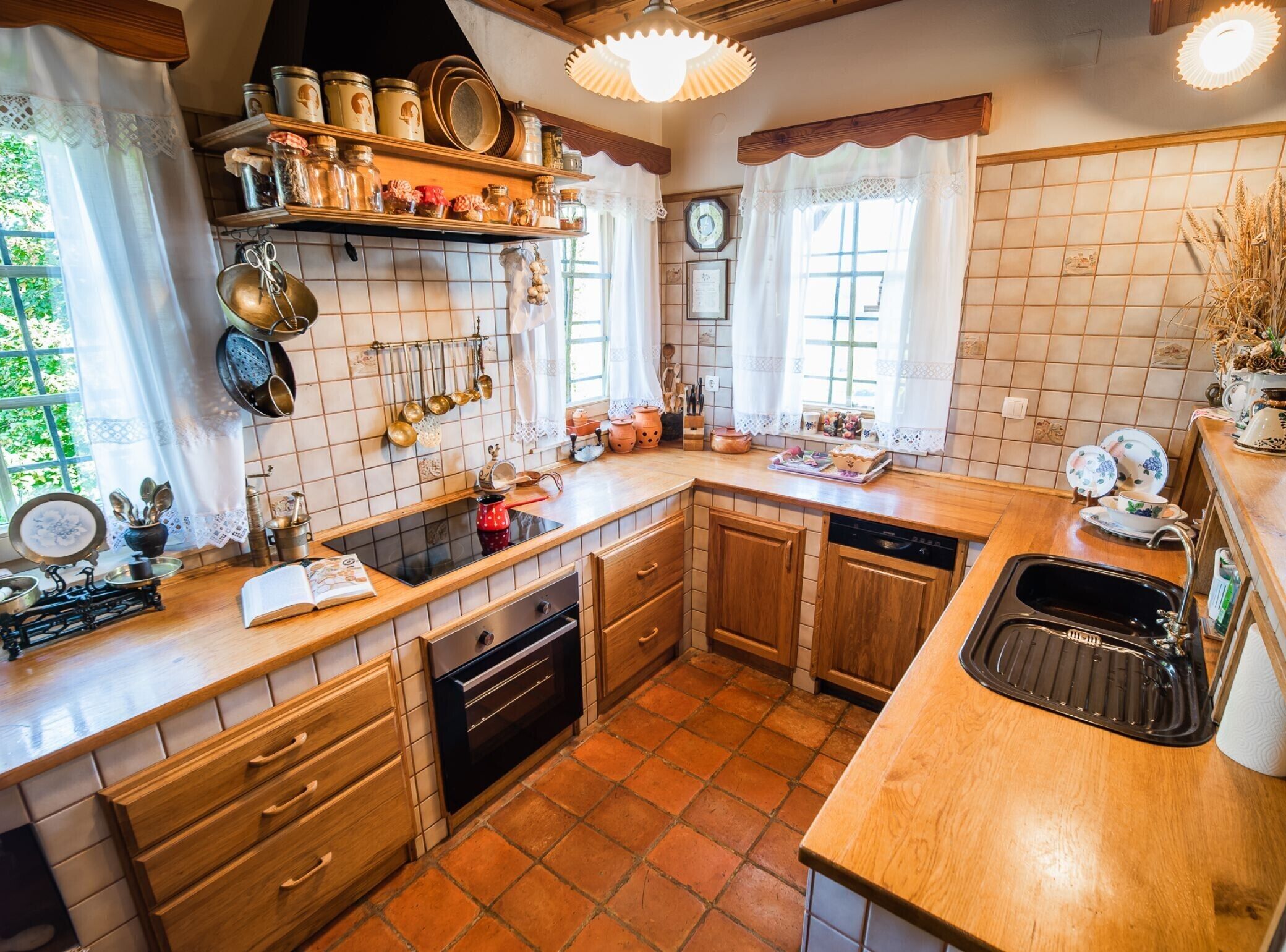 A kitchen with wooden cabinets, a black countertop, and a tiled backsplash. There are shelves with jars and kitchen utensils, and a window with curtains.