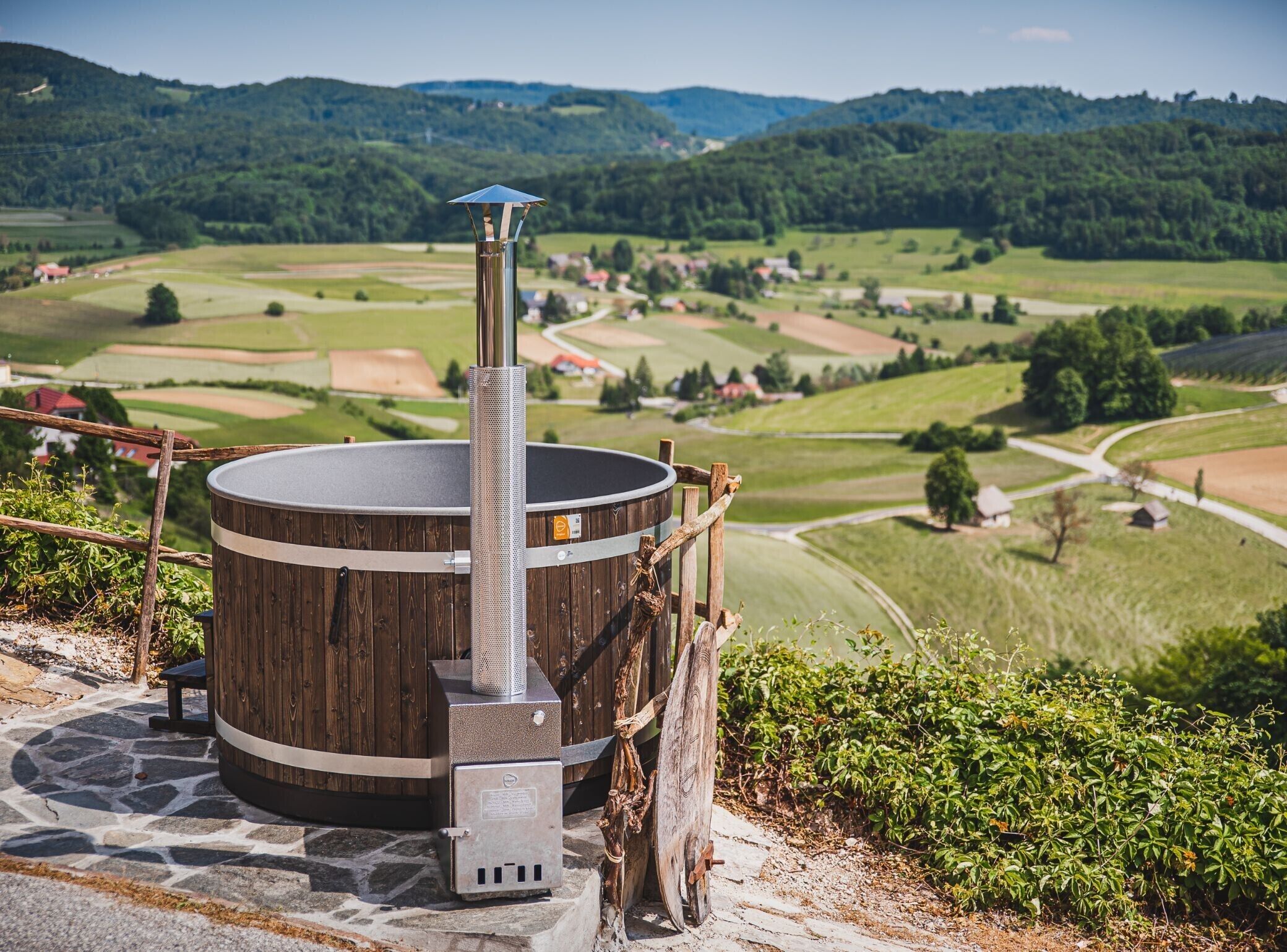 Un sauna rustique installé à l’extérieur, avec une base en forme de tonneau en bois, un conduit de cheminée en métal et une vue imprenable sur une vallée verdoyante.