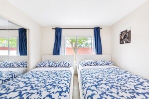 A room with two beds, a window with curtains, a wall-mounted picture, and a view of greenery outside.